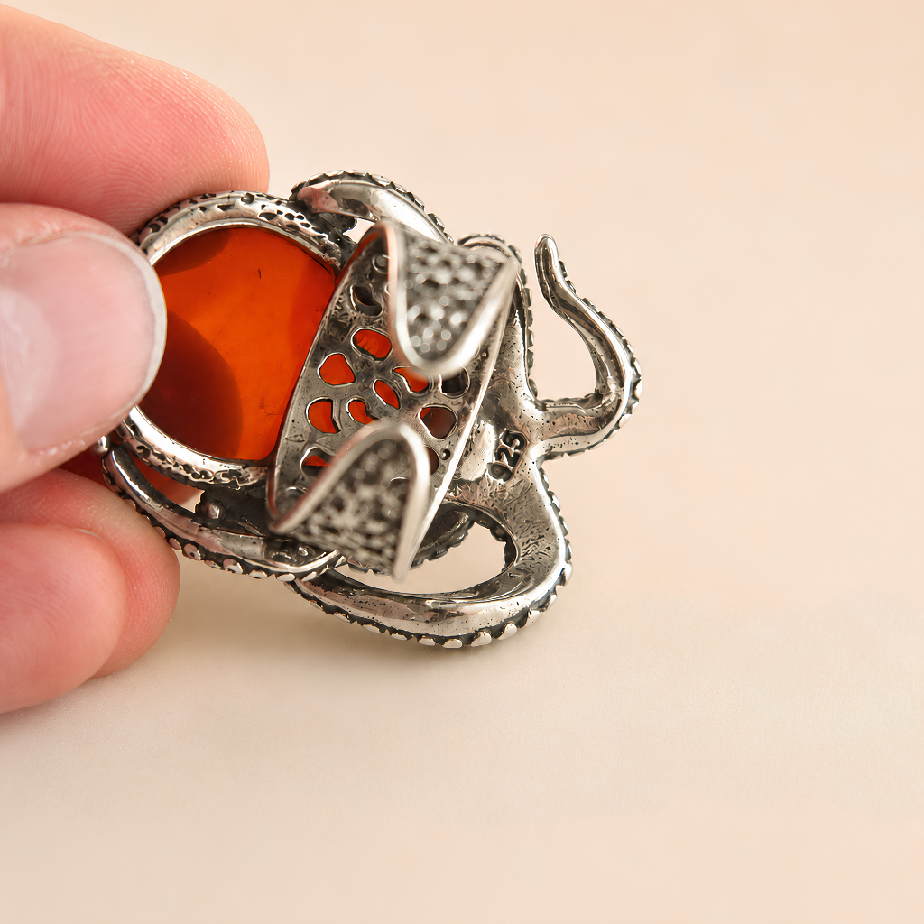 A close-up of a hand holding a silver-octopus ring with a vibrant orange-red stone in a heart-shaped setting, against a solid pale beige background.