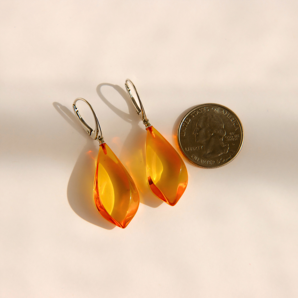 A pair of teardrop-shaped Baltic amber drop earrings with silver leverback hooks, shown next to a US quarter coin for scale on a warm off-white background.