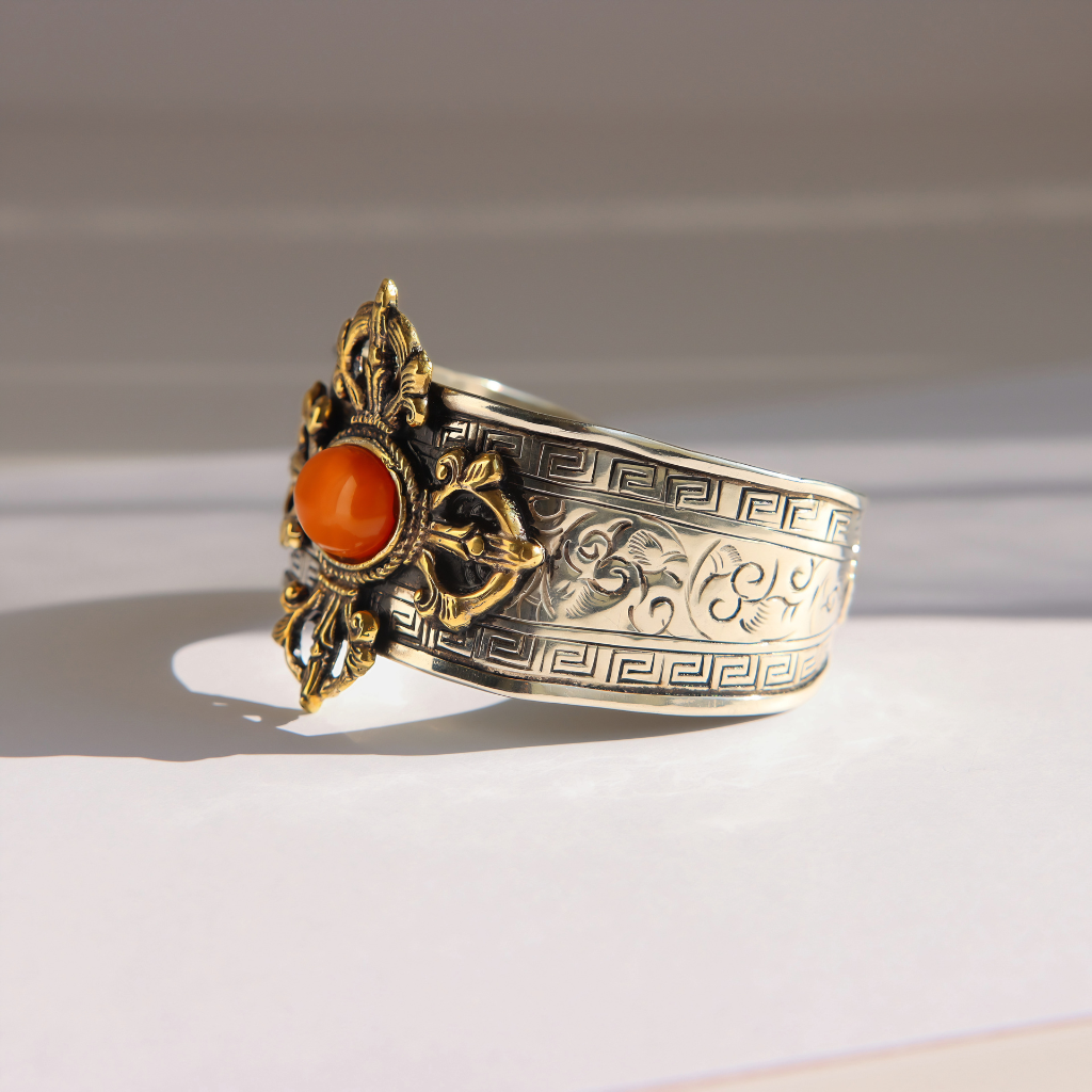 An ornate silver amber bracelet with engraved patterns, a gold-toned filigree setting, and a round orange cabochon gemstone, photographed on a light gray surface with soft shadows.