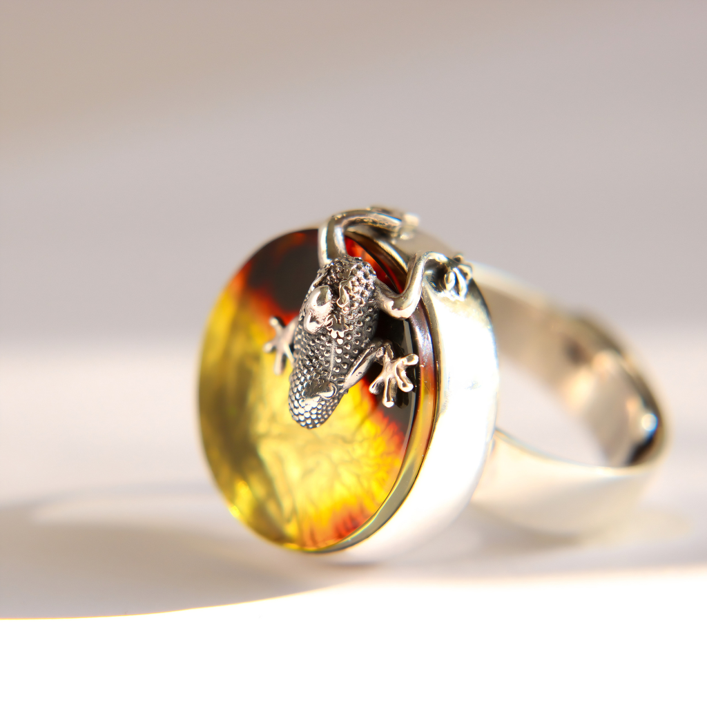 A silver amber ring featuring a round amber cabochon with a yellow and orange marbled pattern and a detailed silver frog climbing on the front, set against a clean white background.
