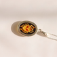A silver oval pendant amber scarab necklace featuring a black background with a stylized yellow and orange beetle design, shown on a light beige surface with a soft shadow.