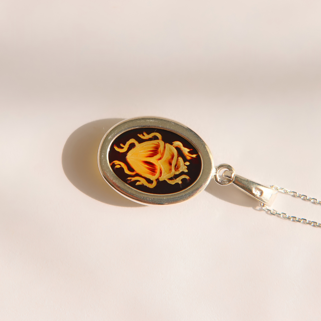 A silver oval pendant amber scarab necklace featuring a black background with a stylized yellow and orange beetle design, shown on a light beige surface with a soft shadow.