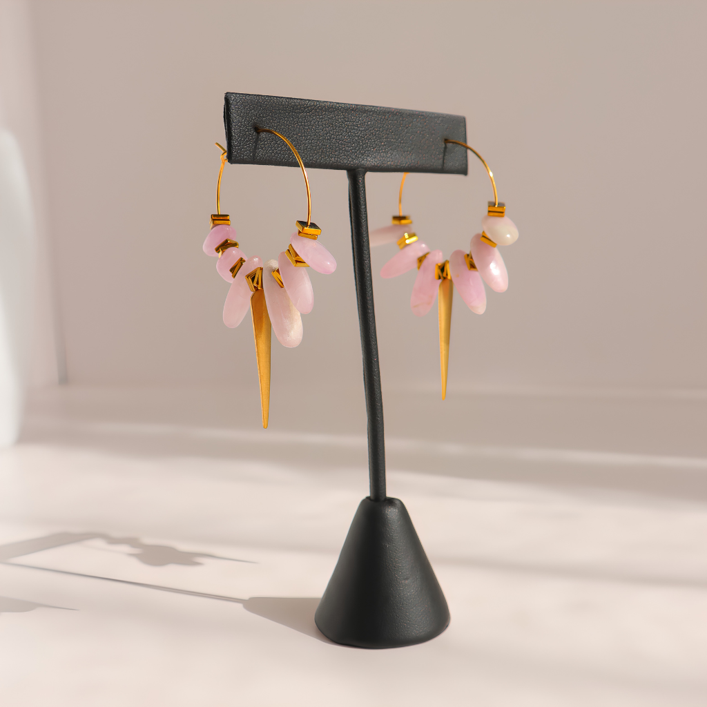 A pair of gold kunzite dagger hoop earrings with dangling pink stone clusters and teardrop charms, displayed on a black jewelry stand against a soft beige background.