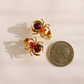 A pair of gold spider-shaped earrings with amber-red centers, shown next to a US quarter coin for scale on a light beige background.