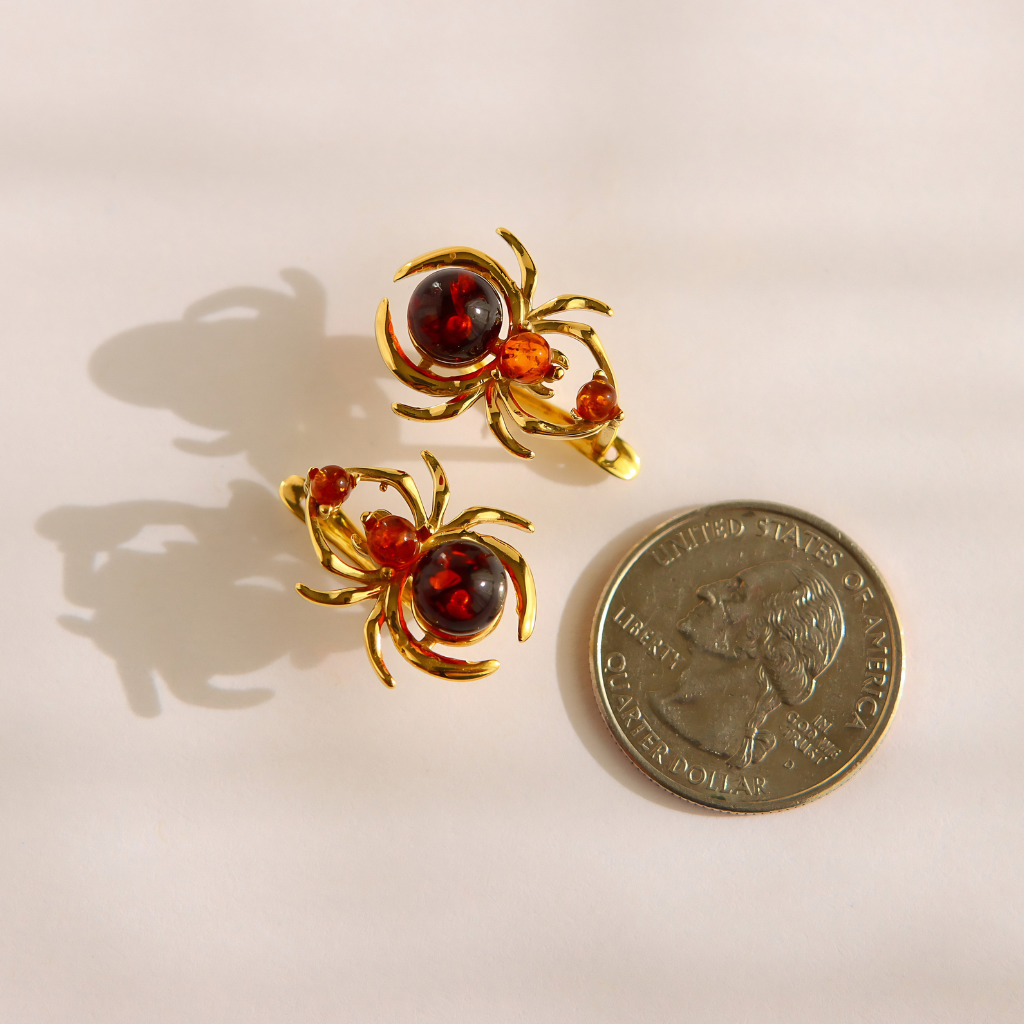 A pair of gold spider-shaped earrings with amber-red centers, shown next to a US quarter coin for scale on a light beige background.