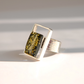 A silver amber ring featuring a rectangular green stone with gold flecks, set in a geometric silver frame with a textured silver band, photographed on a white surface with soft shadows.