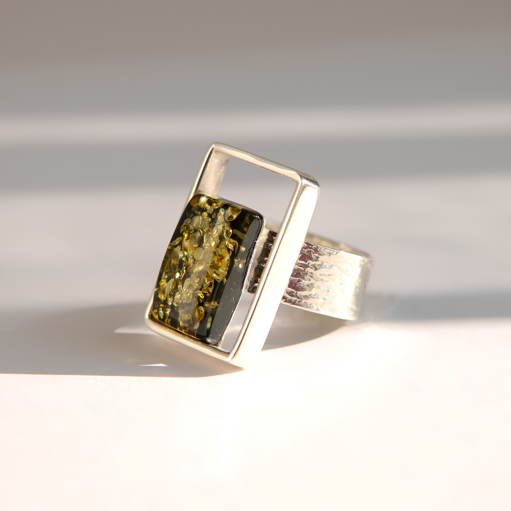 A silver amber ring featuring a rectangular green stone with gold flecks, set in a geometric silver frame with a textured silver band, photographed on a white surface with soft shadows.