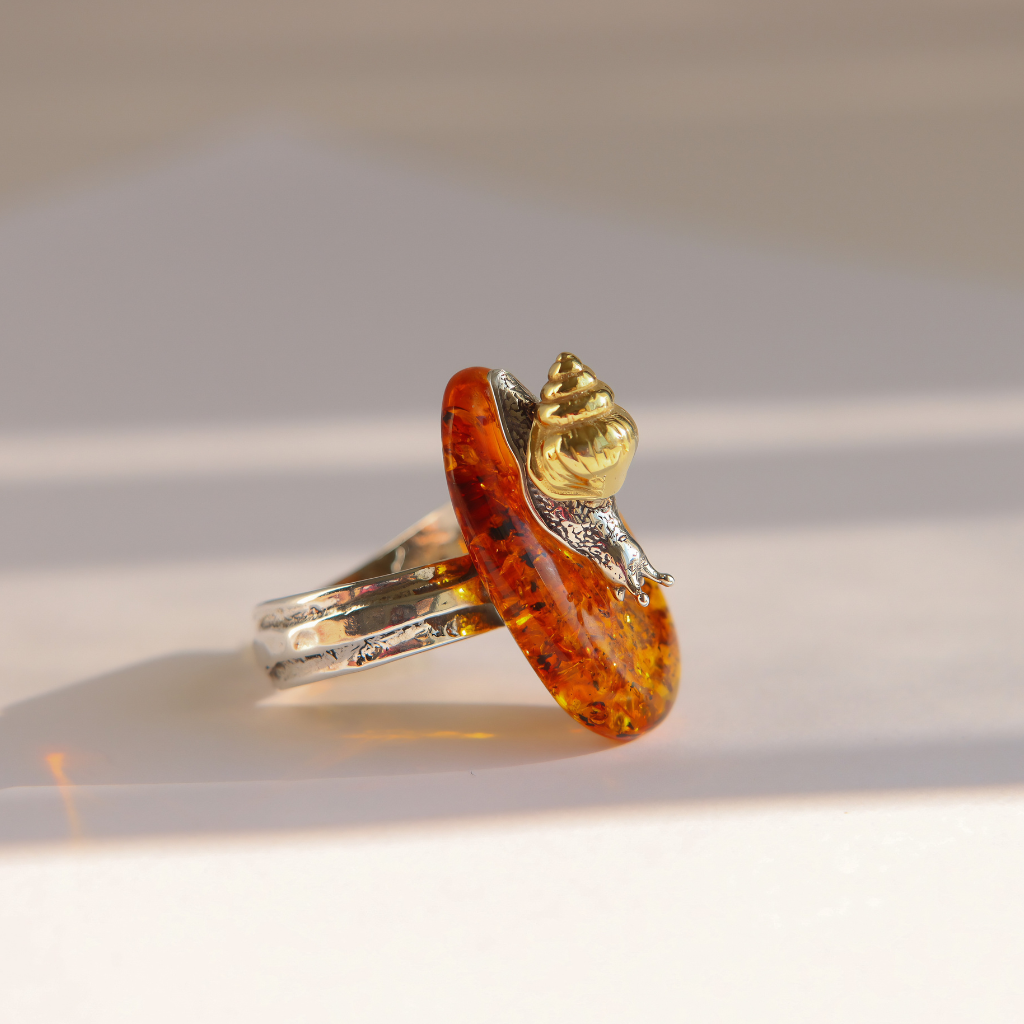 A handmade amber ring featuring a large, polished orange-brown amber gemstone with tiny inclusions, set on a silver band with a small gold snail accent, photographed on a white surface with soft shadows.