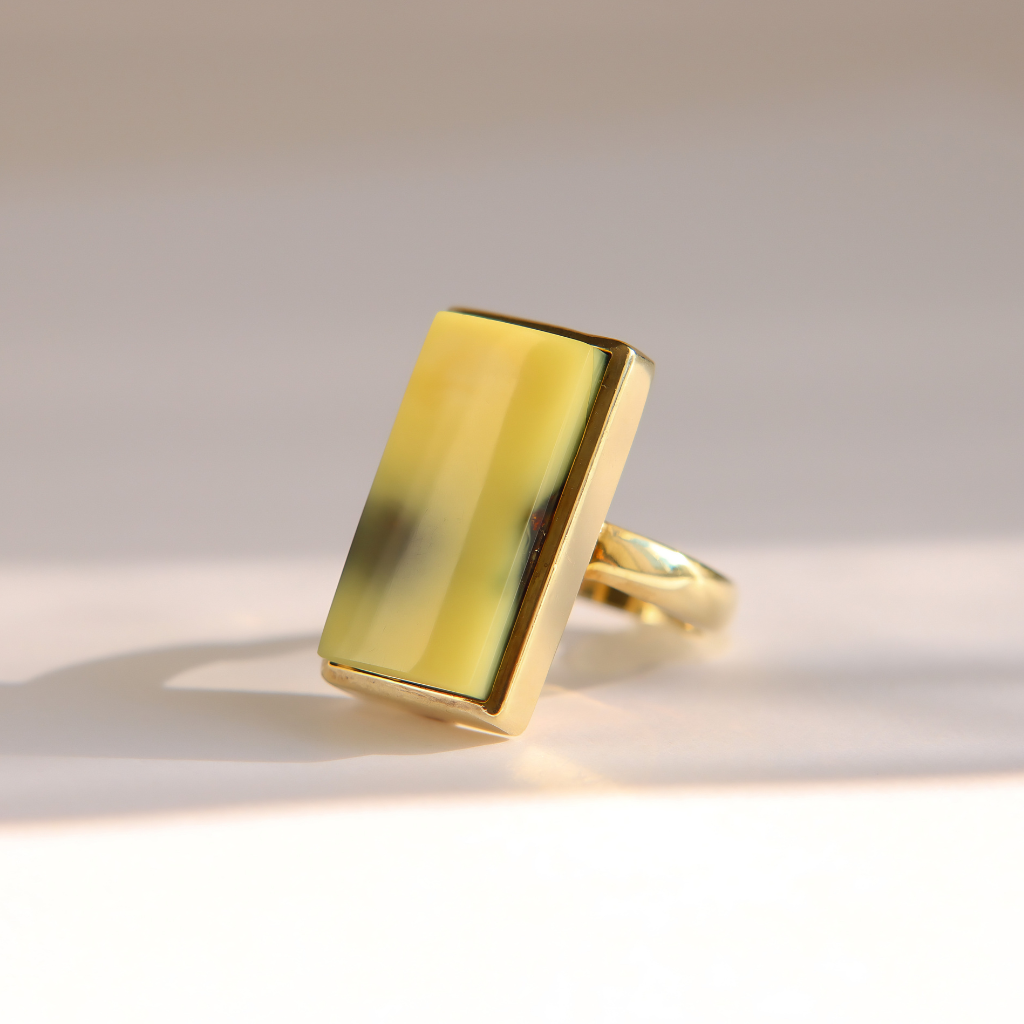 A gold Baltic amber ring featuring a large, polished rectangular yellow-green stone with a glossy finish, set against a clean, light beige background.