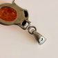 A close-up of a silver-toned frog pendant necklace featuring a small oval amber-orange inset and a dangling teardrop charm, shown on a plain light background.