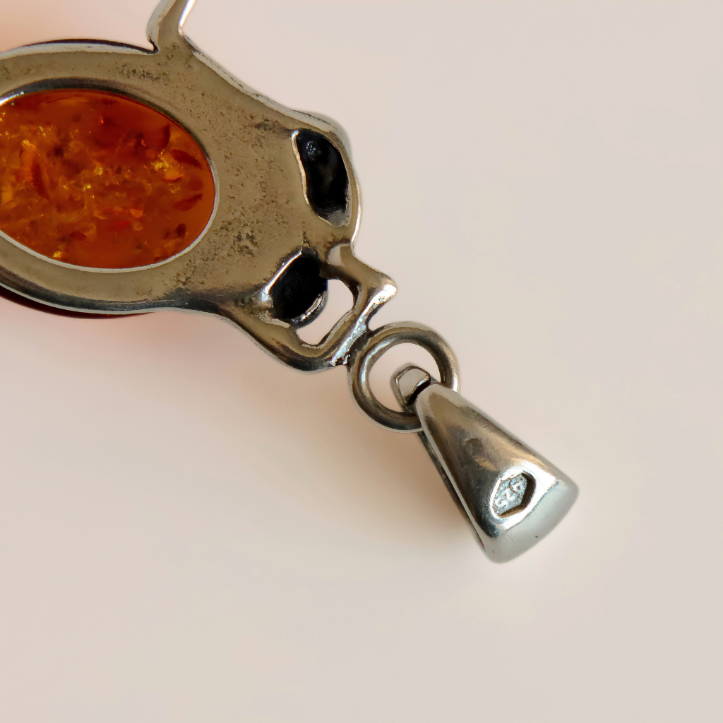 A close-up of a silver-toned frog pendant necklace featuring a small oval amber-orange inset and a dangling teardrop charm, shown on a plain light background.
