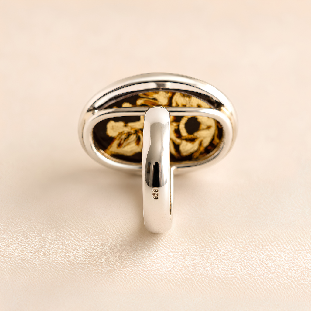 A silver-toned amber cameo ring with an oval face featuring a black and gold floral pattern, photographed on a light beige textured background.