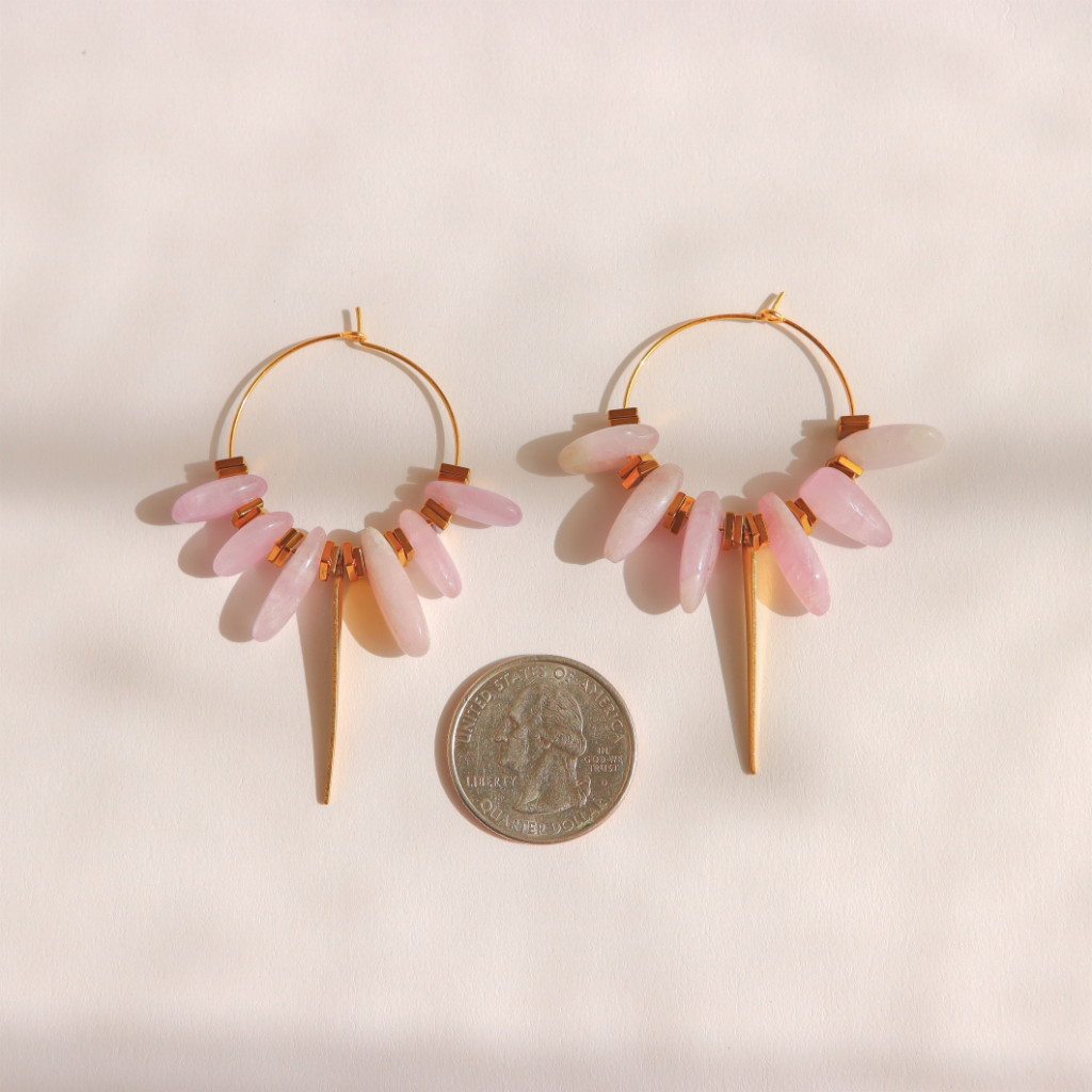A pair of gold kunzite hoop earrings with dangling light pink stone beads, displayed on a warm off-white background with a US quarter coin below for scale.
