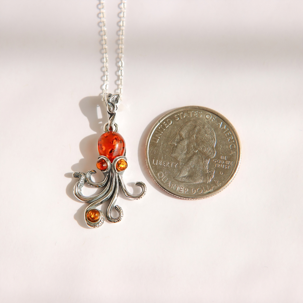 A silver amber octopus pendant necklace with three amber-orange gemstones, shown next to a US quarter coin for scale on a white background.