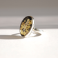 A silver amber cameo ring featuring an oval cabochon stone with a black background and intricate carved floral and butterfly patterns, photographed on a light gray surface with a soft shadow.
