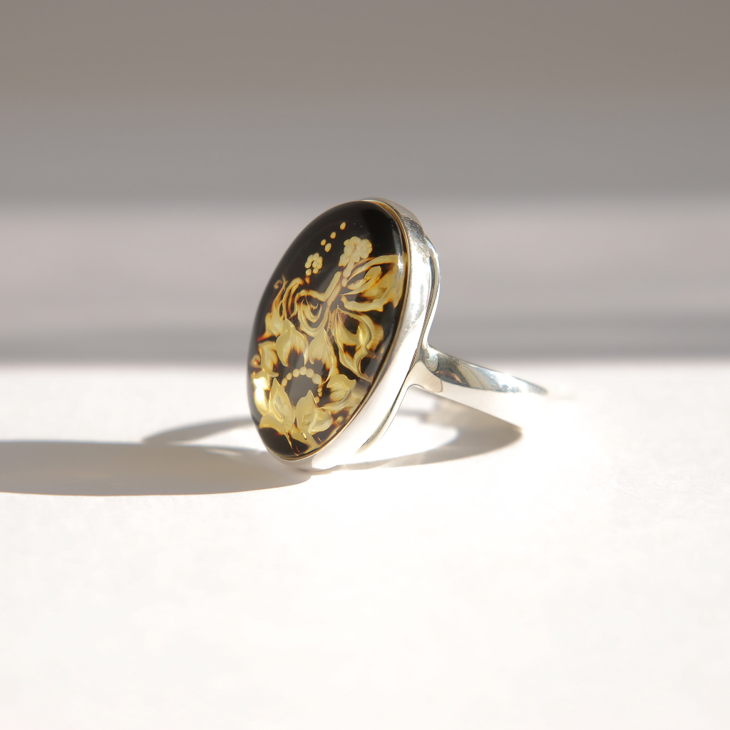 A silver amber cameo ring featuring an oval cabochon stone with a black background and intricate carved floral and butterfly patterns, photographed on a light gray surface with a soft shadow.