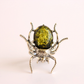 A unique green amber beetle ring featuring a large, oval, olive-green amber cabochon with gold flecks in the center, mounted on a silver-toned spider body and legs.