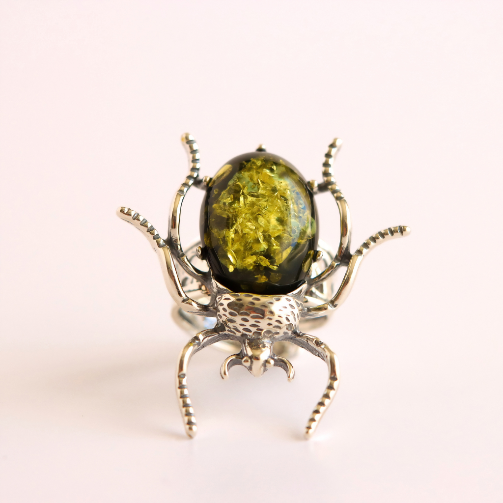 A unique green amber beetle ring featuring a large, oval, olive-green amber cabochon with gold flecks in the center, mounted on a silver-toned spider body and legs.