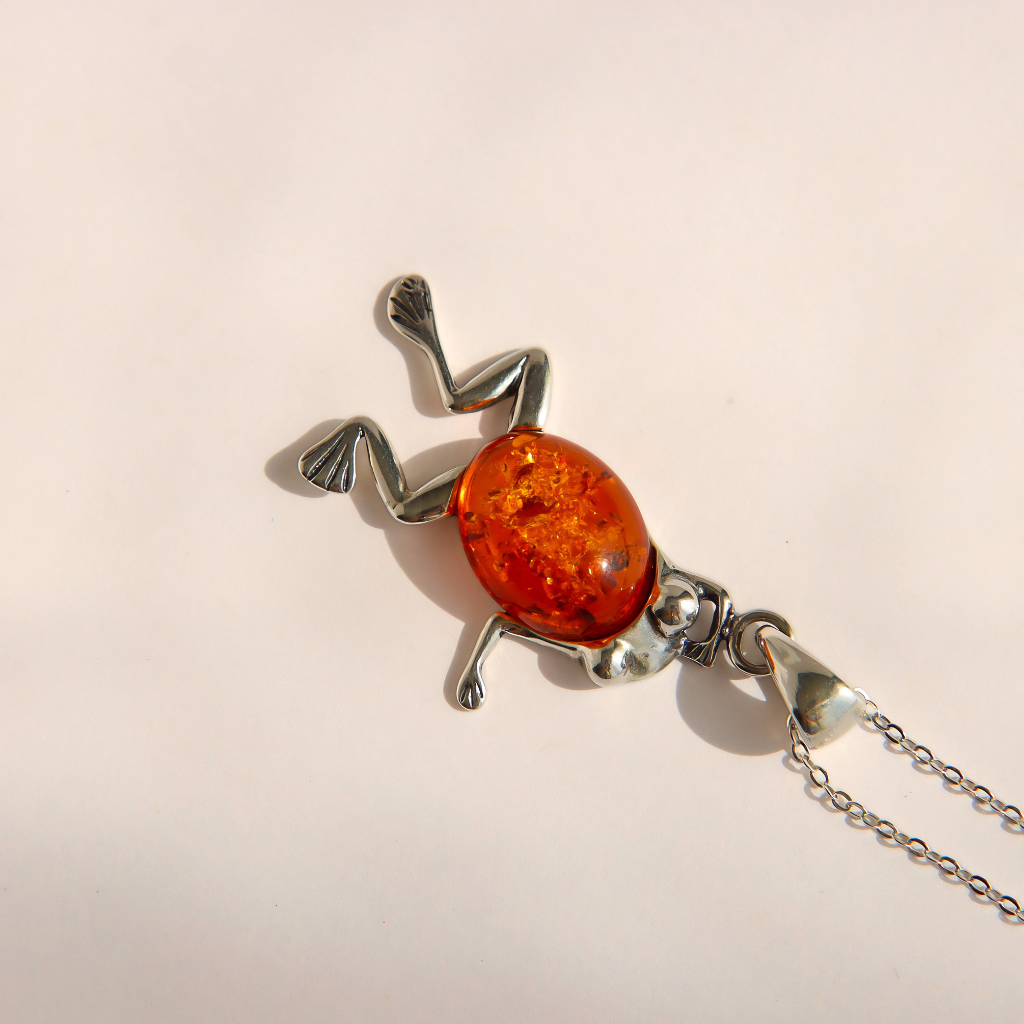 A amber frog pendant necklace made of polished silver with a central amber-orange oval centerpiece, shown on a plain light background with part of the chain visible.