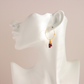 A gold hoop earring with a dangling cluster of small red gemstones, displayed on a white female mannequin head against a soft pink background.
