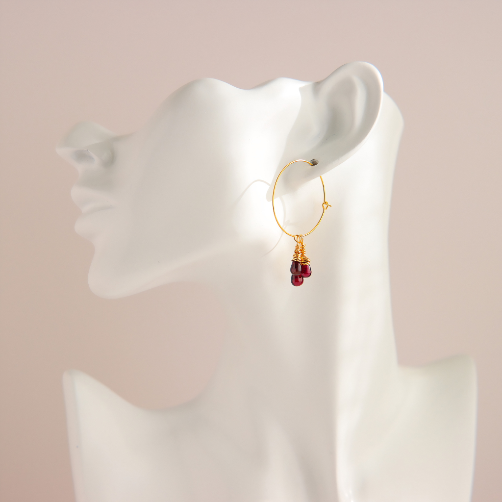 A gold hoop earring with a dangling cluster of small red gemstones, displayed on a white female mannequin head against a soft pink background.