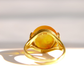 A gold ring featuring a large, round, honey amber stone, photographed against a bright white background with a soft shadow.