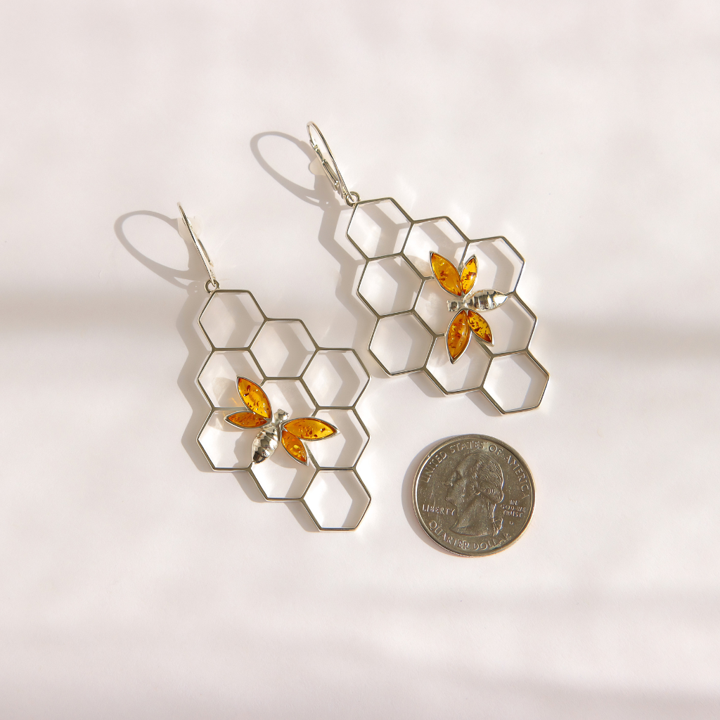 A pair of silver-toned dangle bee earrings with a honeycomb pattern and small amber-orange bee accents, displayed on a white background next to a US quarter for scale.