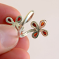 A close-up of a hand holding a Baltic amber flower ring with amber-red inlay details against a soft light pink background showing 925 mark on ring