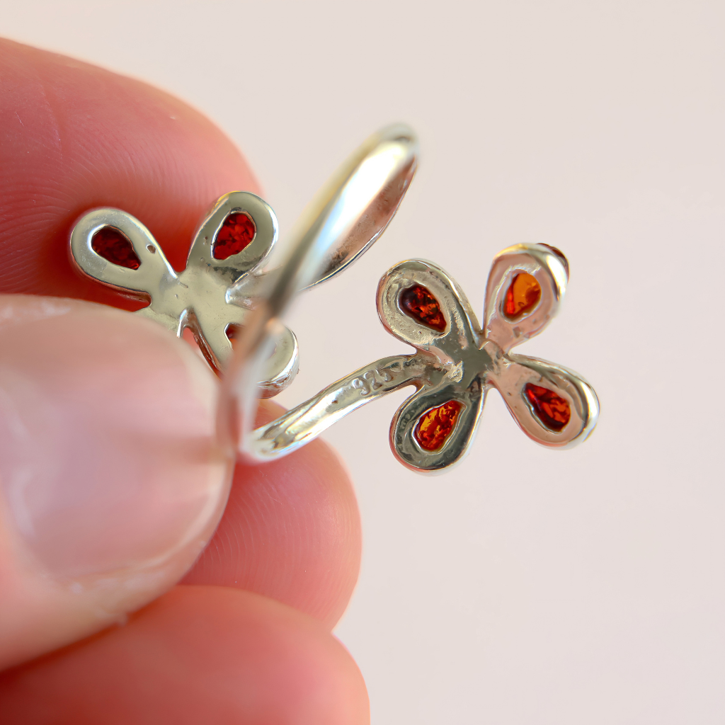 A close-up of a hand holding a Baltic amber flower ring with amber-red inlay details against a soft light pink background showing 925 mark on ring