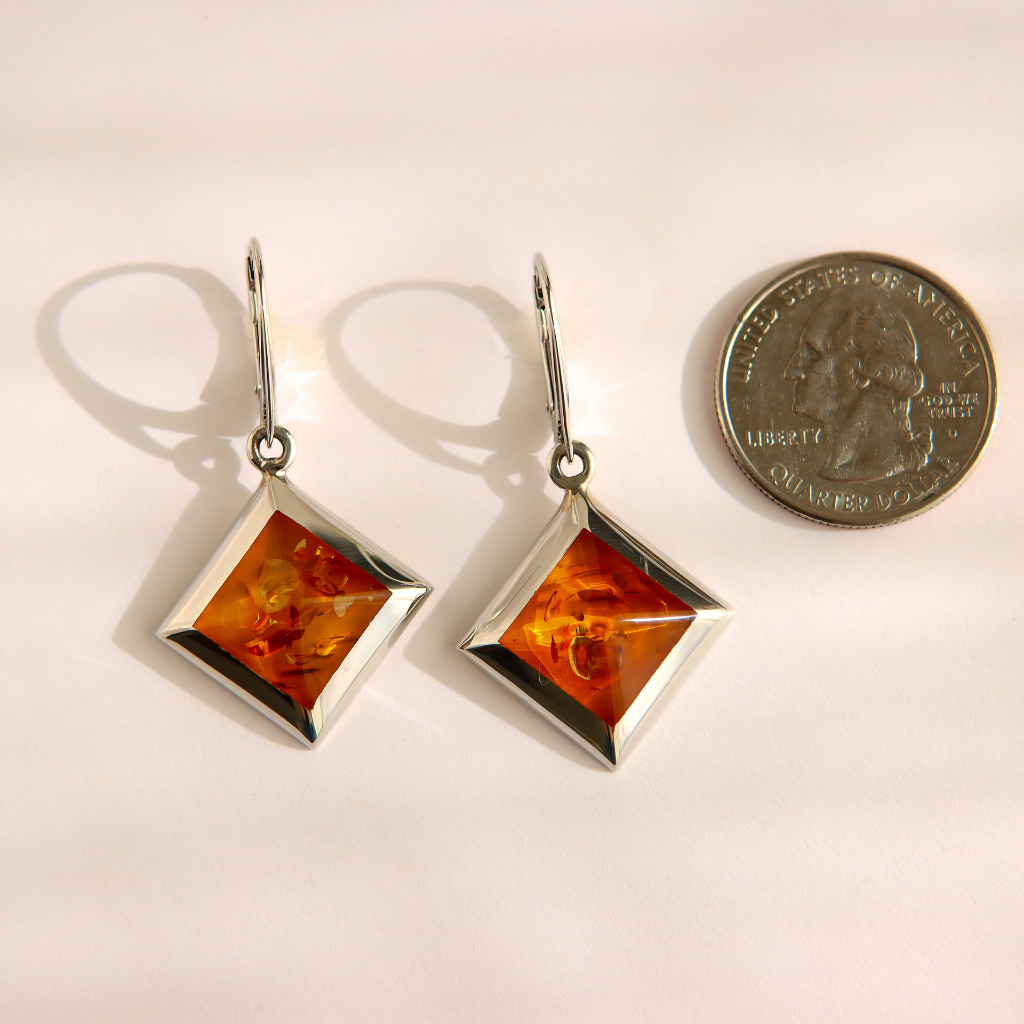 A pair of silver drop amber earrings with square amber stones, shown next to a US quarter coin for scale on a light beige background.