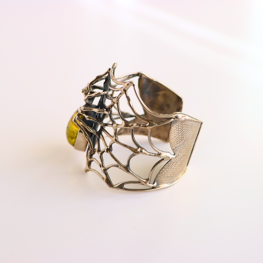A silver metal amber bracelet  with an intricate openwork web-like design and a small round yellow gemstone, photographed on a clean white background.