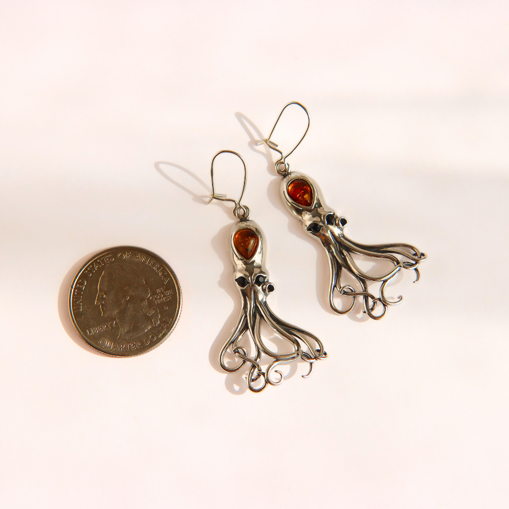 A pair of silver-toned amber octopus dangle earrings with amber-colored gemstones, displayed next to a US quarter coin for scale on a light pink background.