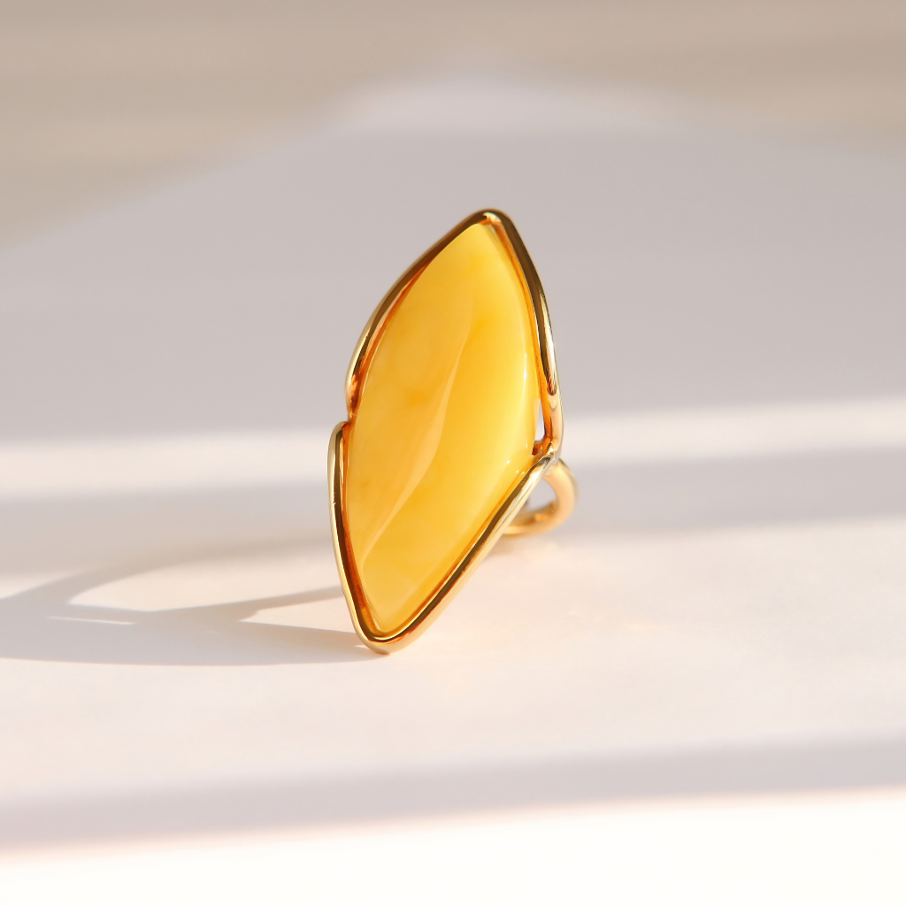 A gold butterscotch amber ring featuring a large, organic-shaped yellow gemstone, photographed on a light beige surface with soft, diagonal shadows.