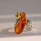 A close-up shot of a amber ring featuring a polished amber oval stone with a small gold and silver snail mounted on top, set against a neutral light gray background.