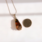 A teardrop-shaped green amber pendant necklace with a gold-toned filigree-wrapped bottom, shown next to a US quarter coin for scale on a white background with soft shadows.