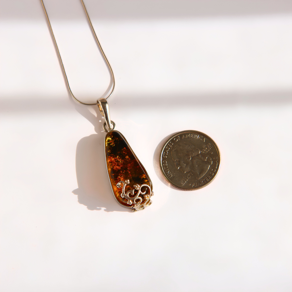A teardrop-shaped green amber pendant necklace with a gold-toned filigree-wrapped bottom, shown next to a US quarter coin for scale on a white background with soft shadows.