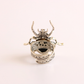 A sterling silver amber beetle ring with intricate filigree detailing and a dark central stone, photographed on a clean, light pink background.