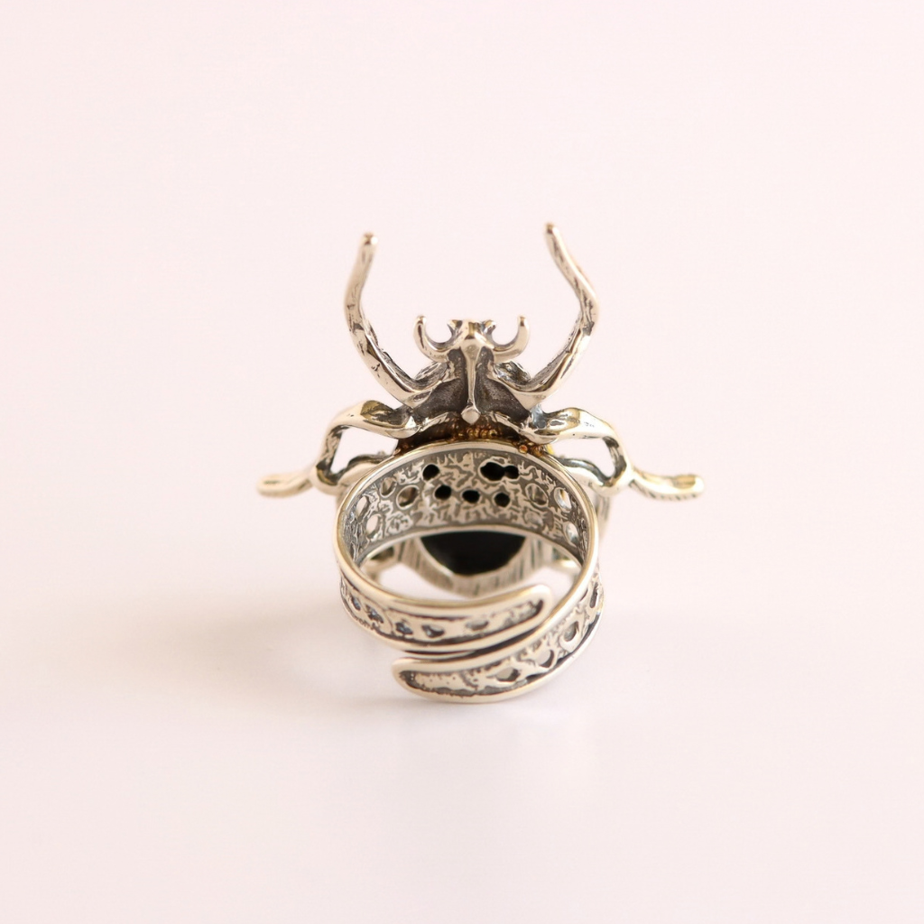 A sterling silver amber beetle ring with intricate filigree detailing and a dark central stone, photographed on a clean, light pink background.