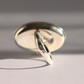 A polished sterling silver amber poison ring shown at an angle on a light gray surface with strong directional shadows.