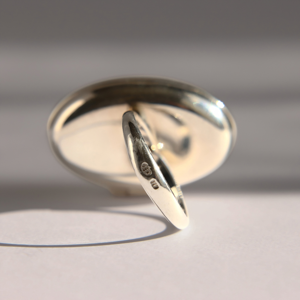 A polished sterling silver amber poison ring shown at an angle on a light gray surface with strong directional shadows.
