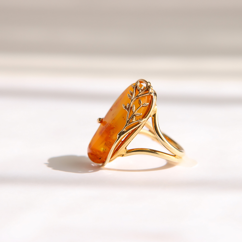 A gold amber ring featuring a large, polished orange-brown stone with delicate gold branch-like detailing, set against a bright, softly shadowed white background.