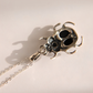 A silver-toned  amber necklace with metal pendant shaped like a stylized beetle or scarab, hanging from a fine chain against a warm off-white background. The pendant features dark oxidized recesses that create black shadows, and is cast with a sharp shadow to the left.