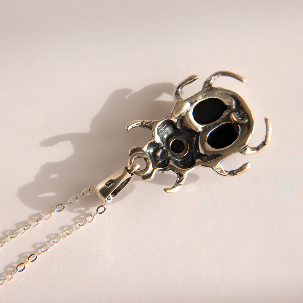 A silver-toned  amber necklace with metal pendant shaped like a stylized beetle or scarab, hanging from a fine chain against a warm off-white background. The pendant features dark oxidized recesses that create black shadows, and is cast with a sharp shadow to the left.