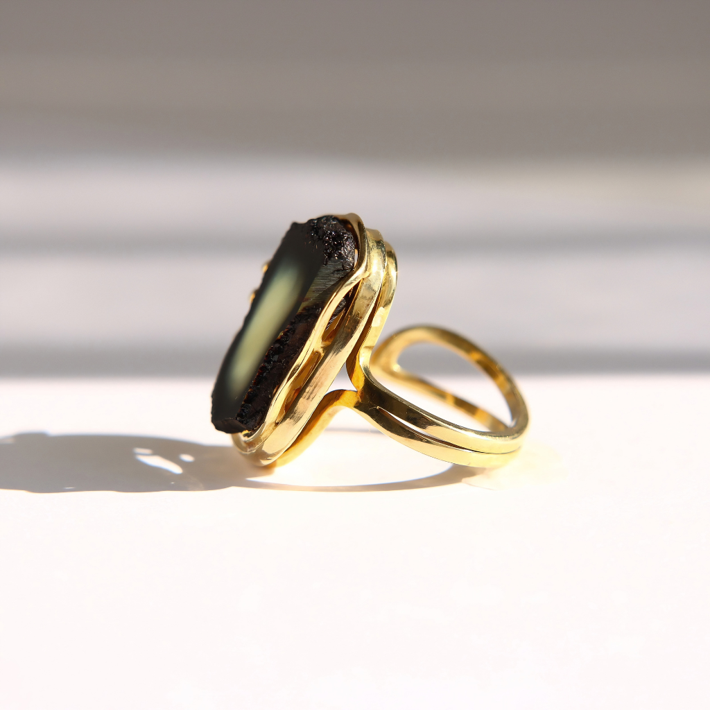A gold amber ring featuring a large, irregular dark green to black stone, photographed on a white surface with soft shadows and bright light.