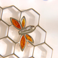 A close-up of a silver bee-shaped earring piece with amber-orange inlaid details, resting on a white hexagonal grid surface showing 925 silver stamp.