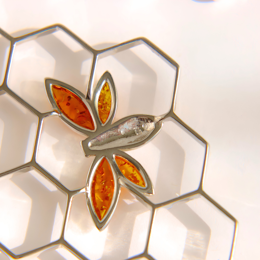 A close-up of a silver bee-shaped earring piece with amber-orange inlaid details, resting on a white hexagonal grid surface showing 925 silver stamp.