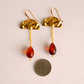 A pair of gold dangle earrings with textured semi-circular tops, short chain links, and teardrop-shaped vintage amber-red Lucite beads, shown next to a US quarter coin for scale on a light beige background.