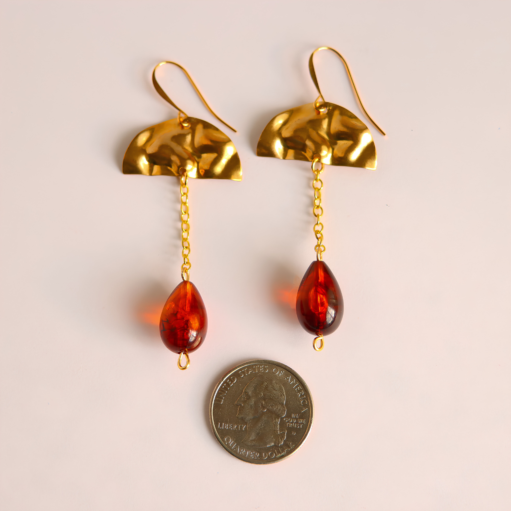 A pair of gold dangle earrings with textured semi-circular tops, short chain links, and teardrop-shaped vintage amber-red Lucite beads, shown next to a US quarter coin for scale on a light beige background.