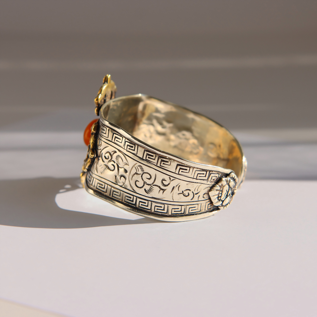 An ornate silver cuff amber bracelet with engraved Greek key and scroll motifs, featuring a small amber gemstone on one side, photographed on a light gray surface with soft shadows.