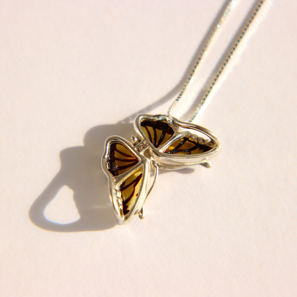 A silver amber butterfly pendant necklace with amber-colored wings, resting on a light cream surface with a soft shadow.